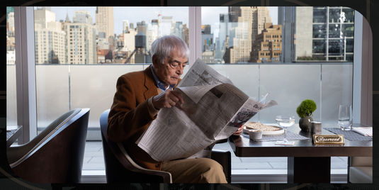 An older man sits alone at a restaurant table by a large window overlooking a city skyline. He is reading a newspaper while a plate of food, a glass of water, and a small vase with greenery sit in front of him. A ‘Reserved’ sign is placed on the table, and the bright daylight highlights the tall buildings outside.