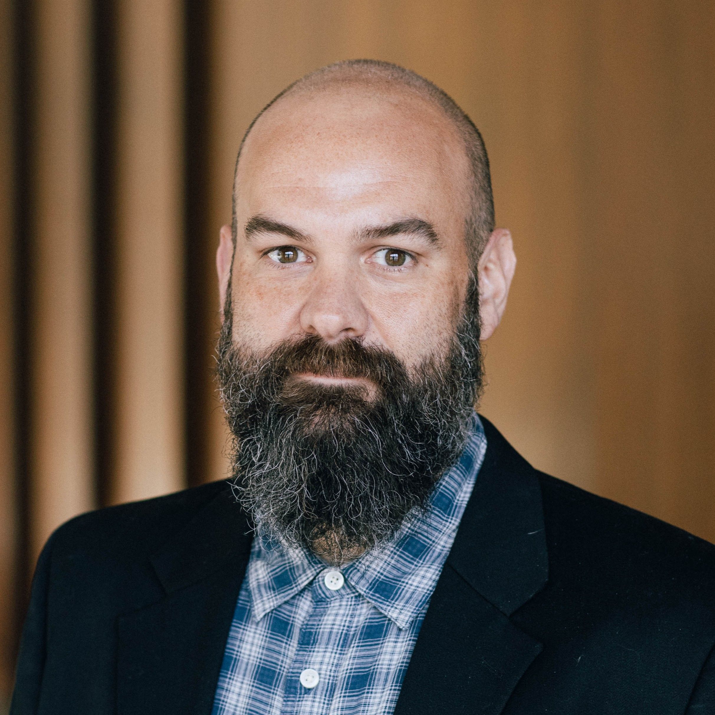 Bald man with a thick beard and mustache wearing a checkered shirt and blazer, standing against a blurred wooden background.