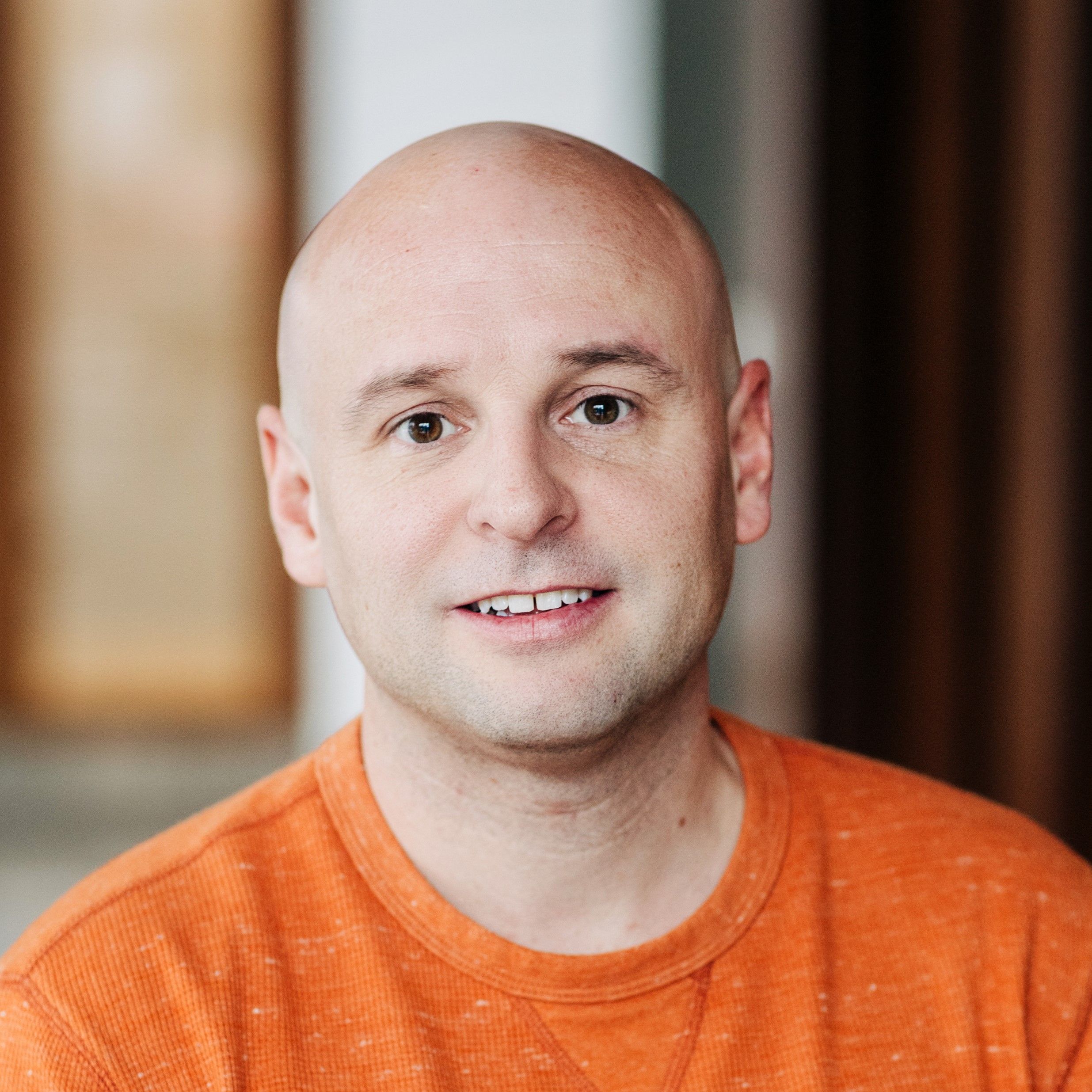 Bald man wearing an orange shirt, smiling gently, with a blurred indoor background.