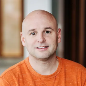 Bald man wearing an orange shirt, smiling gently, with a blurred indoor background.