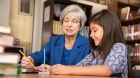 Senior adult teacher, mentor, or tutor assists Latin descent elementary age girl with homework in school classroom or library setting.