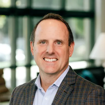 Man in a plaid suit jacket smiling in an office setting with large windows in the background.