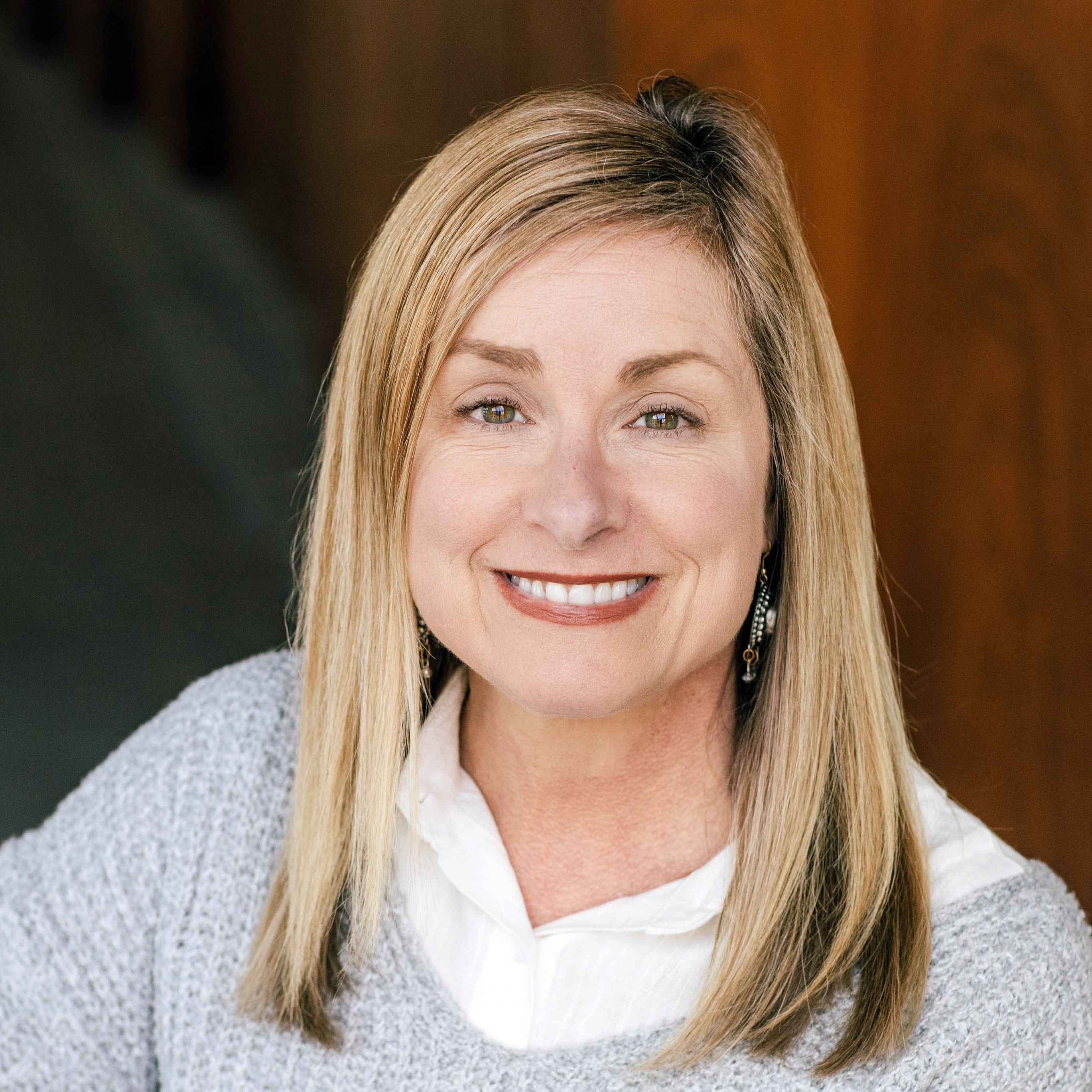 Smiling woman with straight blonde hair, wearing a white blouse and gray sweater, in front of a wooden background.