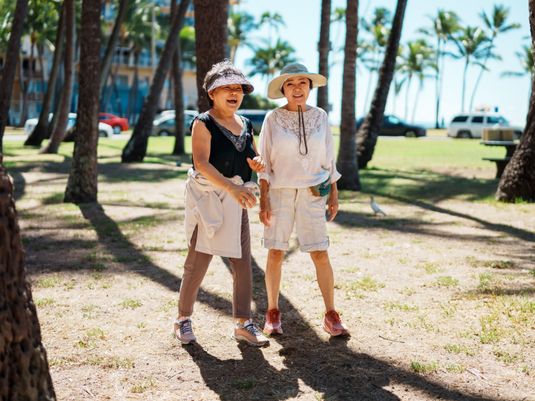 Two women laughing and walking in a sunny park with palm trees, wearing hats and casual summer clothing.