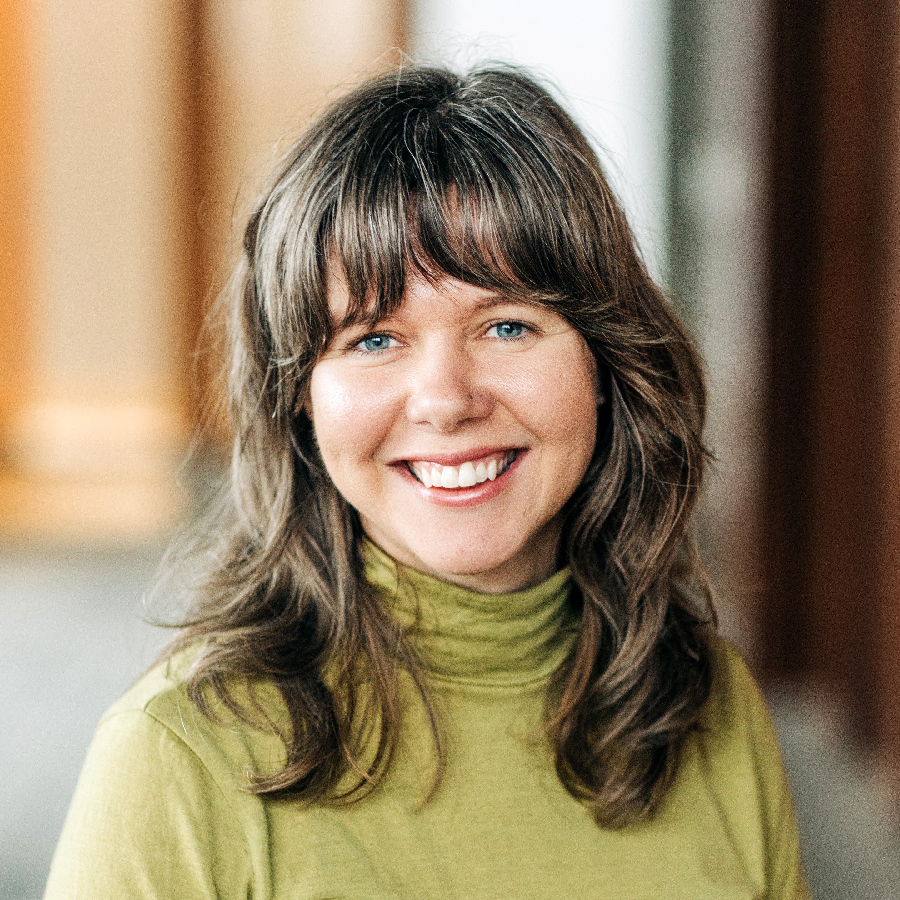 Smiling woman with brown hair wearing a green shirt stands indoors, soft lighting in the background.