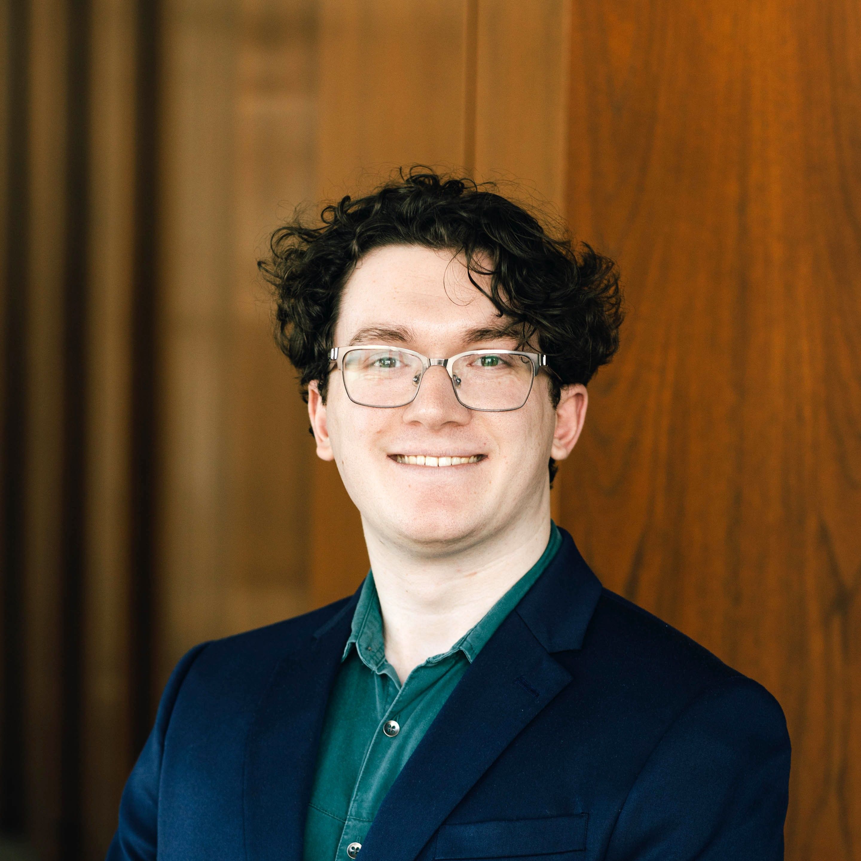 A person with curly dark hair and glasses wearing a blue blazer and green shirt smiles in front of a wooden background.