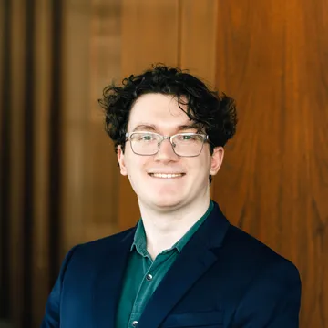 A person with curly dark hair and glasses wearing a blue blazer and green shirt smiles in front of a wooden background.