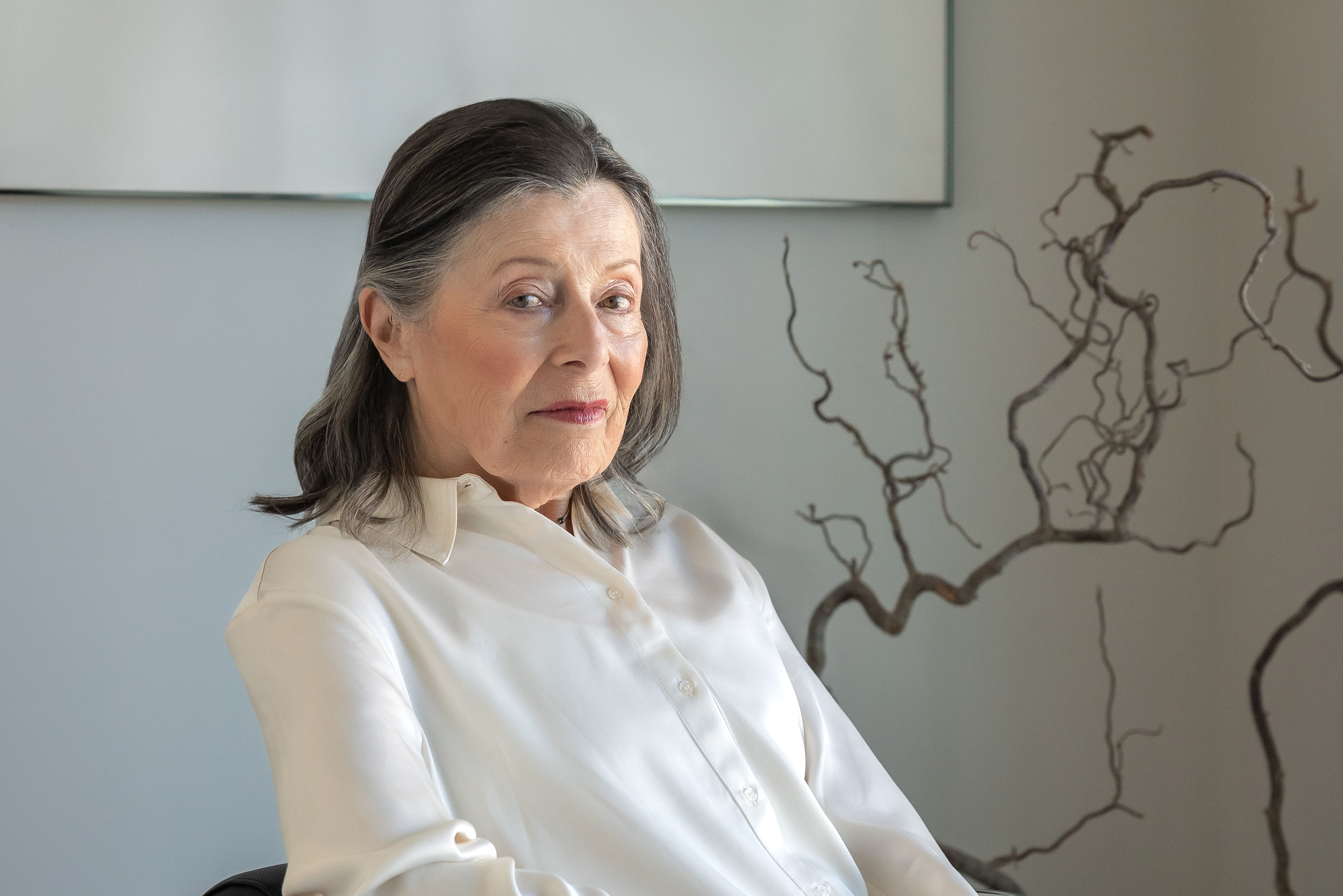 Senior woman with long dark hair, wearing a white blouse, sits in a room with bare branches in the background, looking towards the camera.