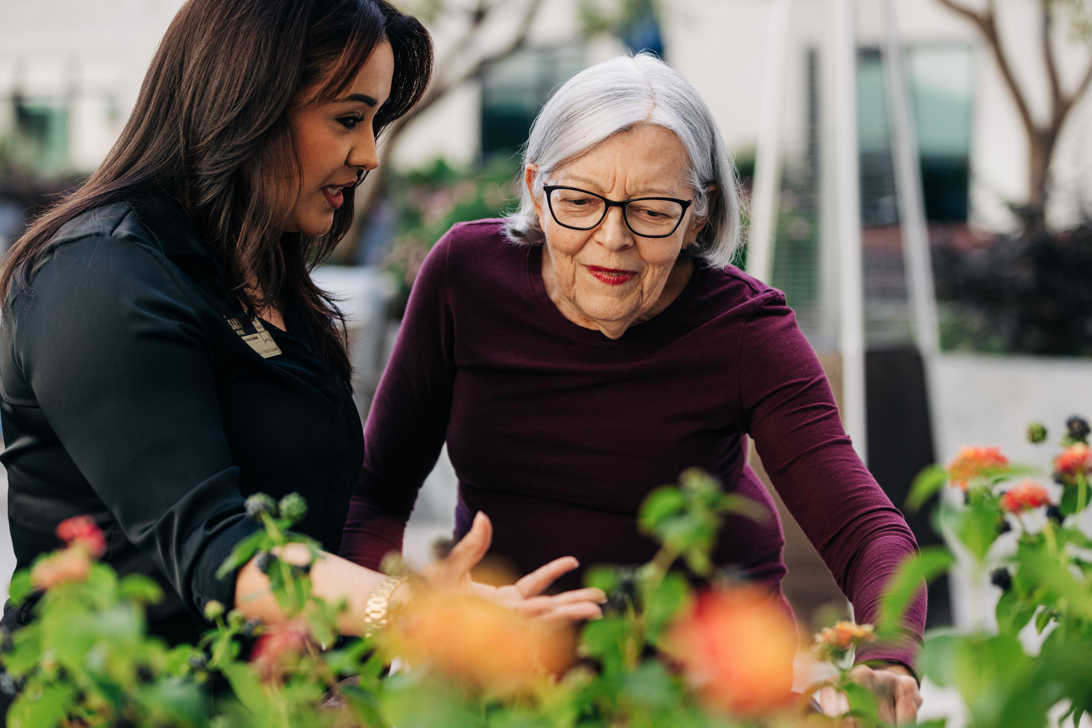 Two women engage in conversation, surrounded by colorful flowers in an outdoor garden setting.