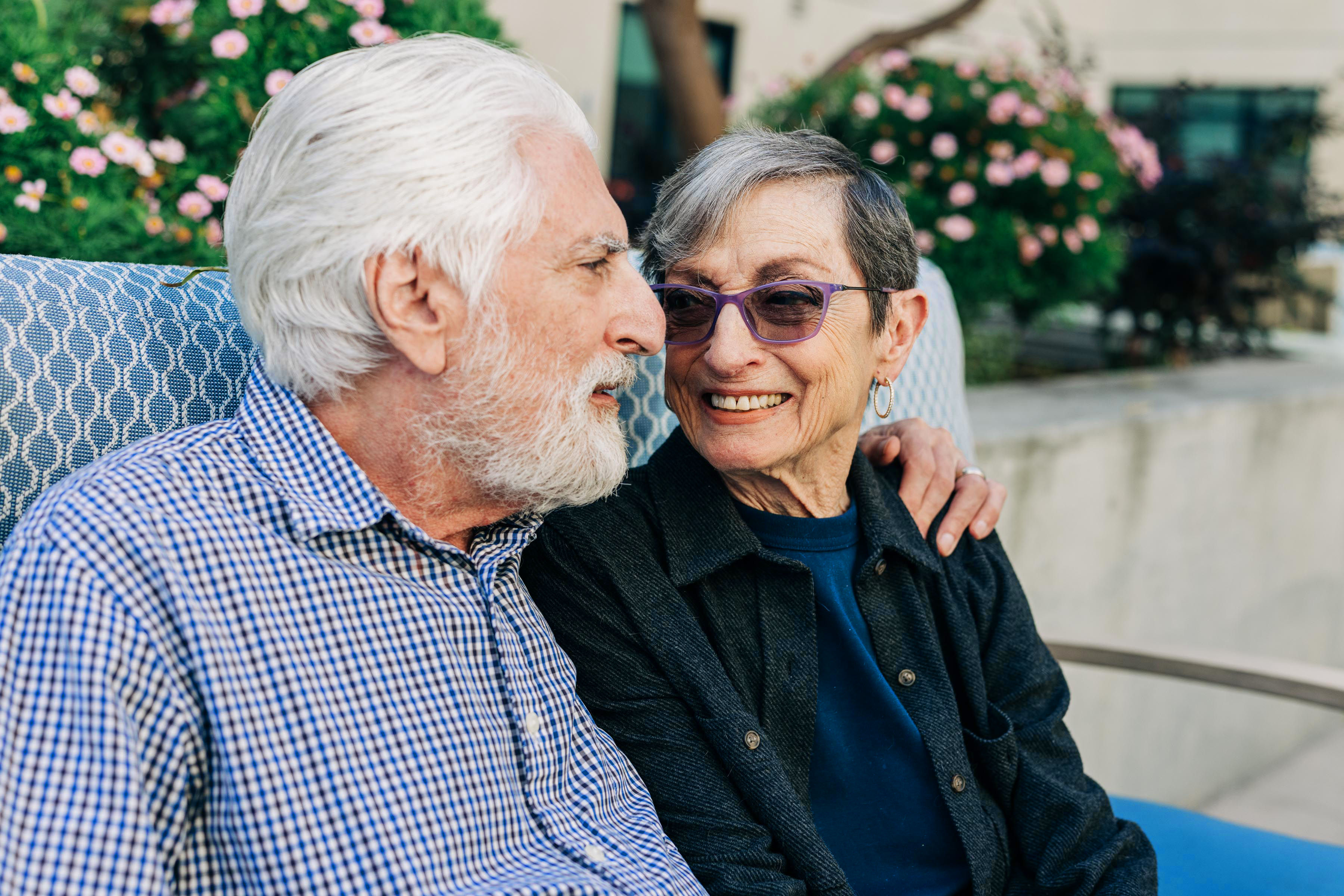 Older couple sitting on a bench, smiling at each other lovingly with flowers in the background.
