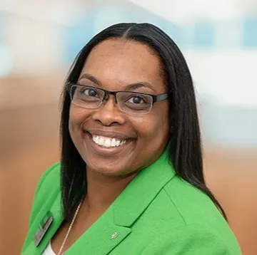 Smiling woman in glasses and a bright green blazer, standing in a softly blurred indoor setting.