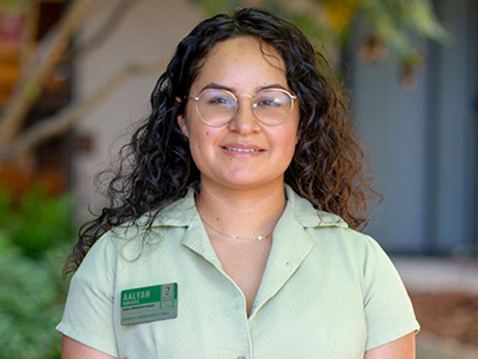 A person with curly hair and glasses, wearing a light green shirt with a name tag, stands outdoors in a garden setting.