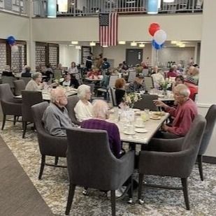 Older people dining in a community hall with festive balloons and an American flag overhead.
