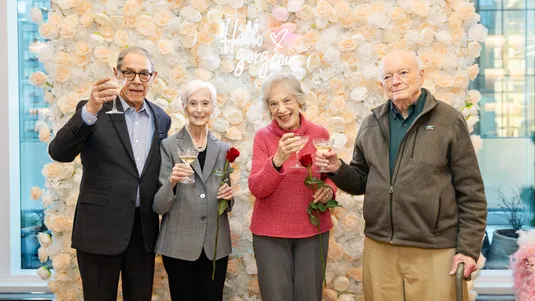 Four elderly people smiling and raising glasses in front of a floral backdrop with a neon "Hello gorgeous" sign. Two hold roses.