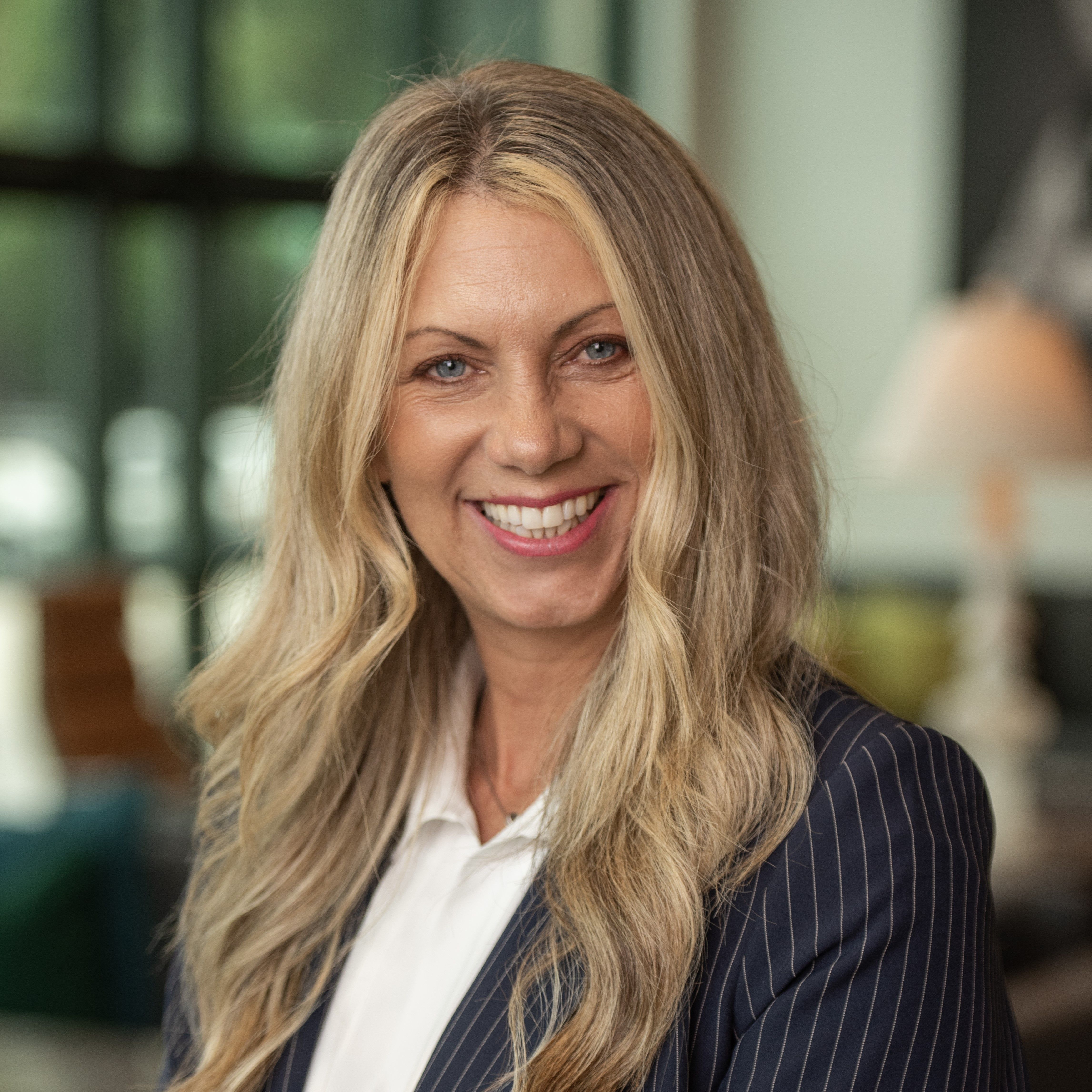 Woman with long blonde hair smiling, wearing a pinstripe blazer over a white shirt in a bright, softly blurred indoor setting.