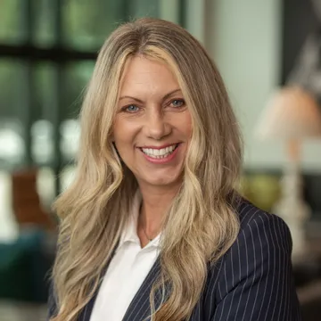 Woman with long blonde hair smiling, wearing a pinstripe blazer over a white shirt in a bright, softly blurred indoor setting.