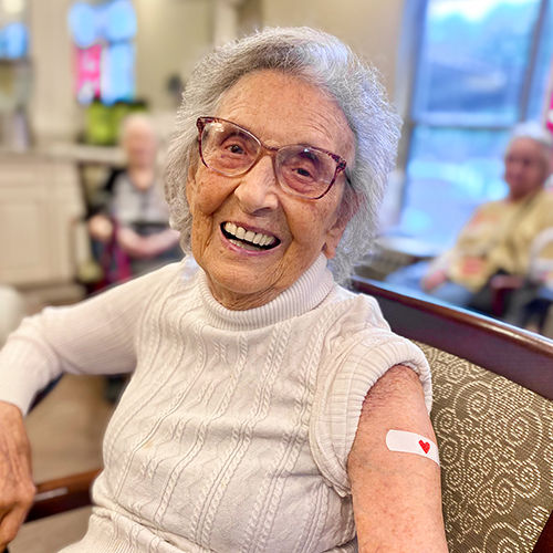 Senior woman smiling, seated indoors, wearing glasses and a white sweater, with a bandage on her arm featuring a red heart.