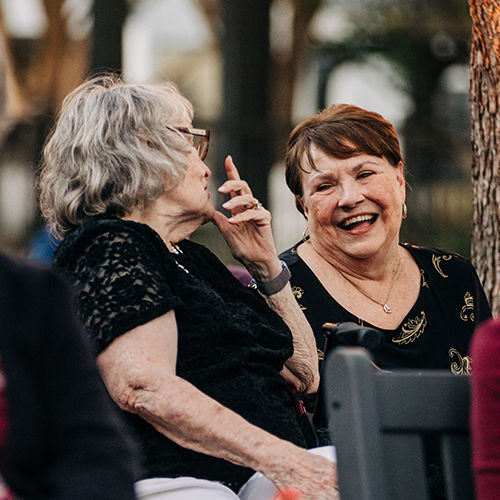 Two senior women sitting on a bench outdoors, smiling and talking. One gestures with her hand, both appear to be enjoying the conversation.