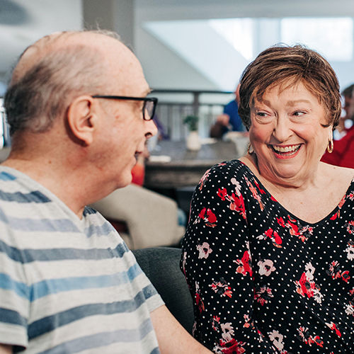 A senior man and woman smiling and talking in a bright room, with the woman wearing a floral blouse and the man in a striped shirt.