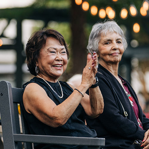 Two senior women sitting outdoors, one clapping and smiling, the other looking ahead. String lights and trees in the background.