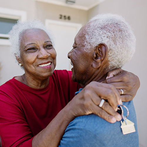Senior couple embracing joyfully outside a house, one holding keys, wearing a red shirt and blue shirt, smiling warmly at each other.