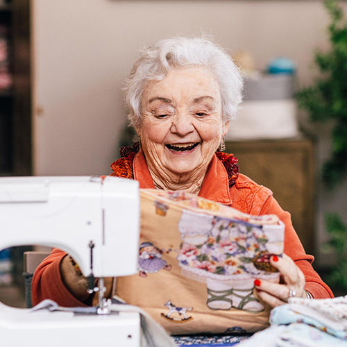 Senior woman smiling while sewing with a machine, holding colorful fabric in a cozy room.
