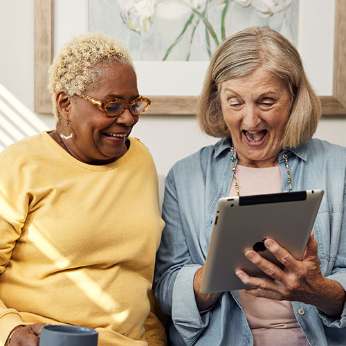 two senior women sitting together on a couch watching an iPad and laughing together