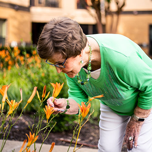 Senior woman in a green sweater leans forward to admire orange flowers in a garden.