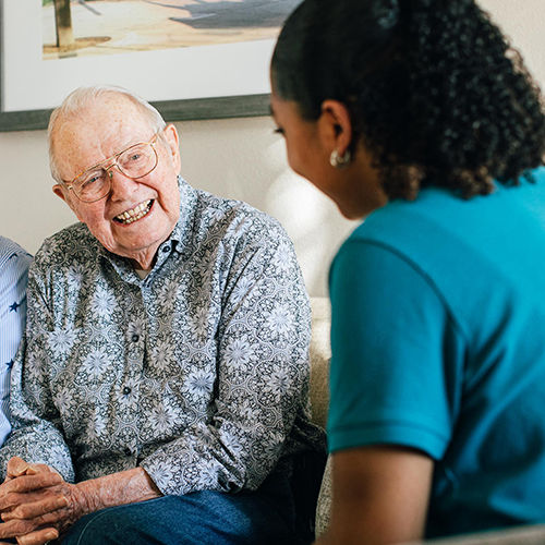 Senior man smiling and talking with a younger woman in a blue shirt, sitting indoors on a couch.
