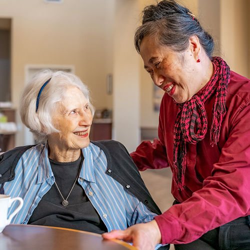 Senior woman sitting and smiling at a woman in a red shirt who is leaning towards her, both appearing happy in a cozy indoor setting.