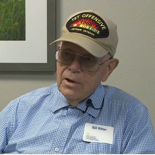 An older man wearing glasses and a "Tet Offensive Vietnam Veteran" cap sits indoors, dressed in a blue checkered shirt.