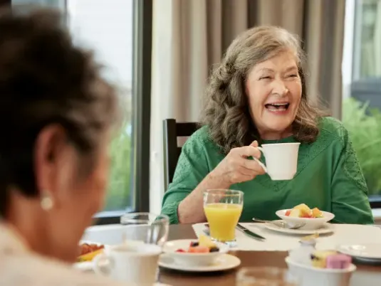 Older woman having breakfast with friends, she is in a green blouse with shoulder length gray hair, and drinking coffee and laughing.
