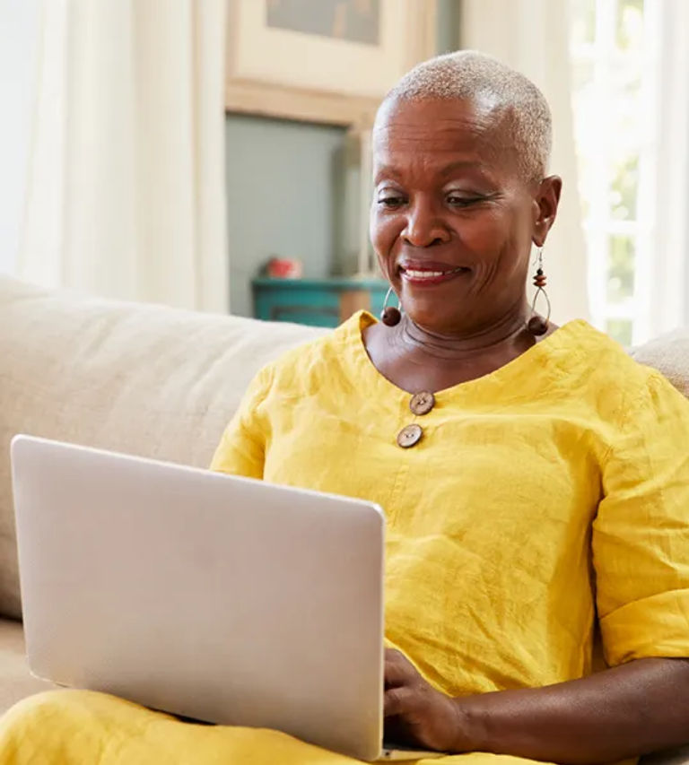 Senior resident in yellow dress sitting on her couch researching senior living options on her laptop.