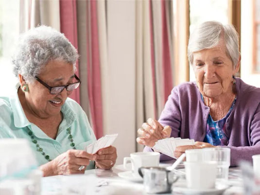 Two female residents smile as they play cards and enjoy refreshing drinks at a table.