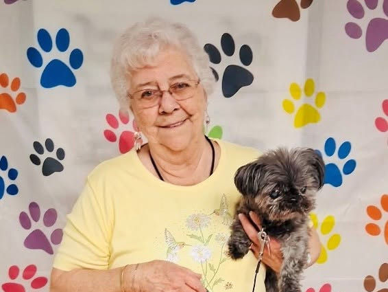 Senior woman in a yellow shirt smiling and holding a small dog, standing in front of a paw print-patterned backdrop.