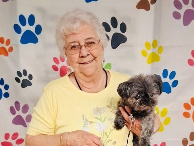 Senior woman in a yellow shirt smiling and holding a small dog, standing in front of a paw print-patterned backdrop.