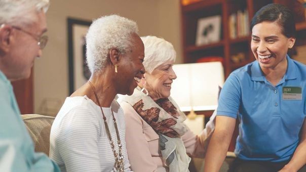 A caregiver in a blue shirt smiles while interacting with three older people sitting together in a cozy room.