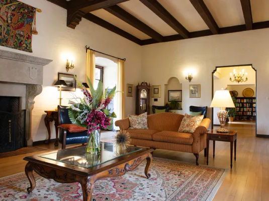 Elegant living room with a striped sofa, floral centerpiece on a glass coffee table, ornate fireplace, and wooden beams on the ceiling.