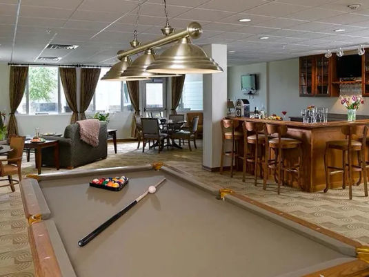 A cozy recreation room with a pool table, bar area, seating, and large windows. Warm lighting and neutral tones create a relaxed atmosphere.