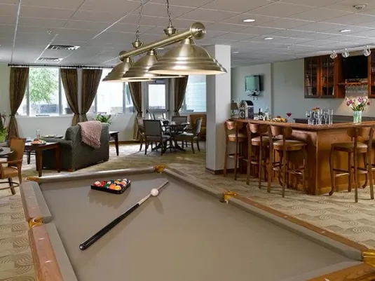 A cozy recreation room with a pool table, bar area, seating, and large windows. Warm lighting and neutral tones create a relaxed atmosphere.