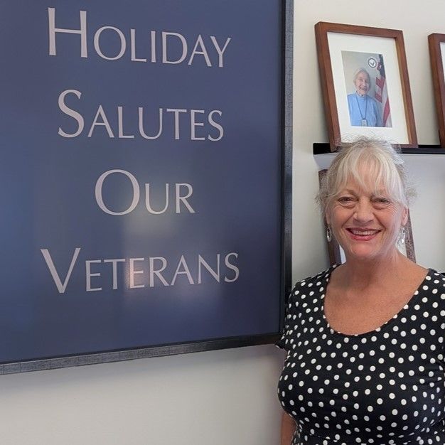 Woman in a polka dot dress stands beside a sign reading "Holiday Salutes Our Veterans." Framed photos are visible in the background.