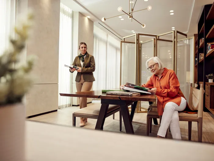 Two women in a modern office; one sits reading at a table, the other stands holding a tablet. Large windows and a bookshelf are visible.