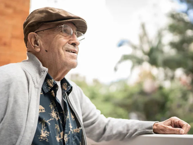 Older man in a cap and glasses, wearing a patterned shirt and cardigan, gazes thoughtfully from a balcony with greenery in the background.