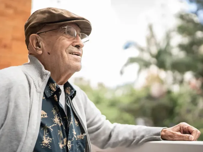 Older man in a cap and glasses, wearing a patterned shirt and cardigan, gazes thoughtfully from a balcony with greenery in the background.