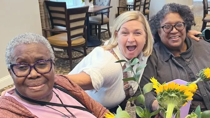 Three women smiling and sitting together at a table with sunflowers, in a cozy room with wooden chairs and patterned carpet.