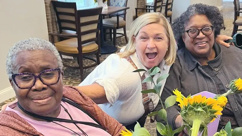 Three women smiling and sitting together at a table with sunflowers, in a cozy room with wooden chairs and patterned carpet.