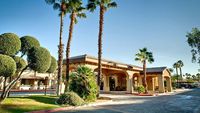 A sunny view of a Mediterranean-style building with arches, surrounded by palm trees and manicured shrubs under a clear blue sky., click to stop animation