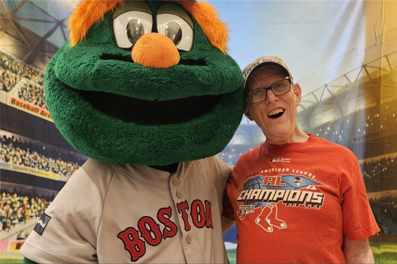 A man in a red shirt poses happily with a large, green, furry mascot wearing a "Boston" jersey, set against a stadium backdrop.