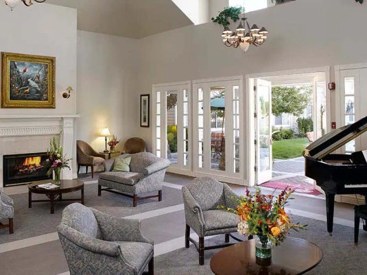Atria Summit Ridge living area with fire place, comfortable searing, grand piano and a wall of doors with windows.