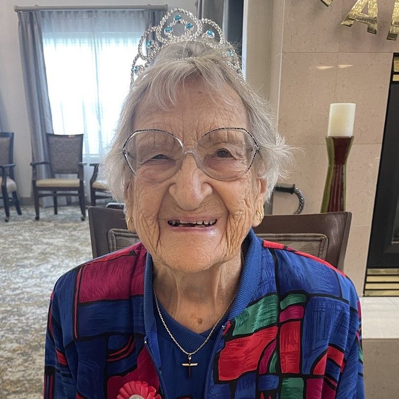 Older woman wearing glasses and a tiara, smiling warmly. She's dressed in a colorful blouse and seated indoors.